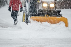 Snow plow clearing a roadway during a winter storm with a worker walking alongside the equipment.
