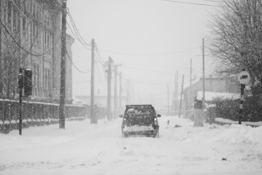 Car driving down a snow-covered road during a heavy winter storm in an urban area.